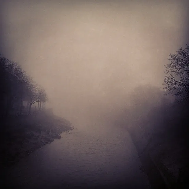 A sepia photo looking directly up a river. It is very foggy. There are trees on either side.