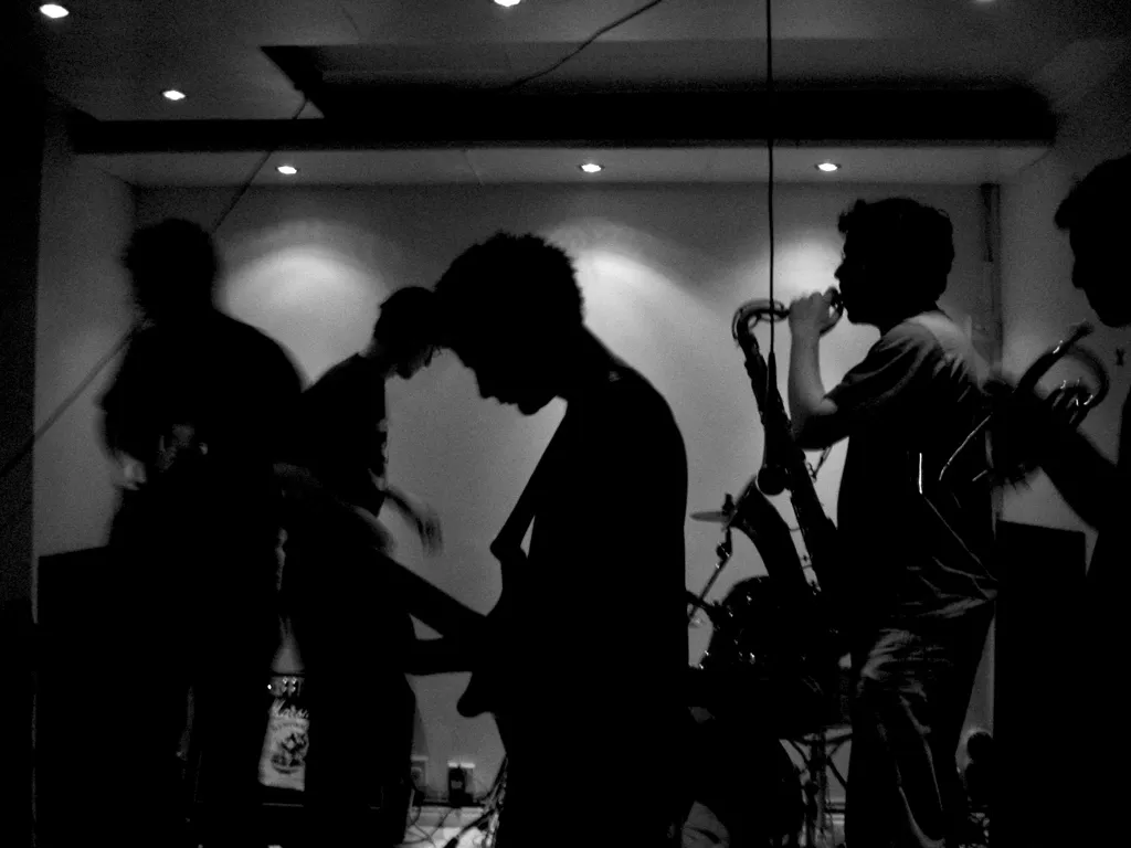 A black and white image of a band. They're mostly in silhouette. A guitarist, bassist, saxophonist and trumpeter are in the foreground. In the background is a drum kit.